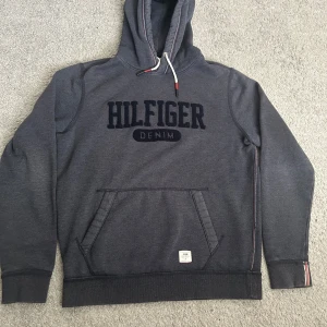 Grå hoodie från Hilfiger Denim - Säljer en snygg grå hoodie från Hilfiger Denim med stor logga på bröstet. Tröjan har en klassisk känguruficka och justerbar huva med snören. Perfekt för en avslappnad stil.