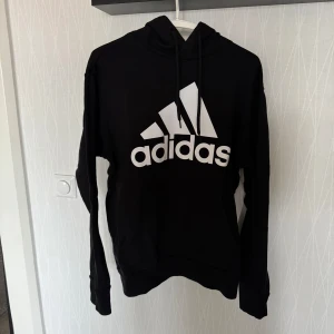 Svart hoodie från Adidas - Säljer en svart hoodie från Adidas med det klassiska logotyptrycket i vitt på framsidan. Tröjan har en justerbar huva med snören och långa ärmar. Perfekt för en sportig och avslappnad stil.
