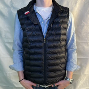 Marinblå Tommy-Hilfiger Väst - Snygg Marinblå dunväst med dragkedja och diskreta detaljer. Perfekt för lager-på-lager under kyliga dagar. Västen har en stilren design med en liten logotyp på bröstet och två praktiska fickor med dragkedja. Storlek S men skulle säga att den passar fint även på M.