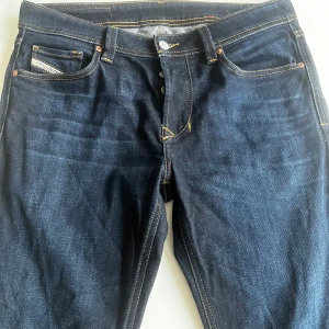Mörkblå jeans från Diesel - Snygga mörkblå jeans från Diesel med klassisk femficksdesign och knappgylf. De har en läderpatch med Diesel-logga baktill och kontrastsömmar som ger en cool look. sitter ganska smalt i pass form. betydligt mörkare färg i verkligheten än på bild 