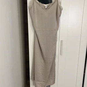 Beige klänning från H&M i satin  - Elegant beige klänning från H&M med spets vid bysten och en subtil slits där bak. Klänningen har en v-ringad design och är tillverkad i ett satin som ger en fin passform. Perfekt för en stilren look.