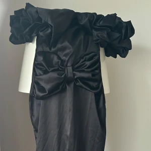 Svart axelbandslös klänning med volangdetaljer - Elegant svart axelbandslös klänning med stora volangdetaljer vid axlarna och en dekorativ rosett framtill. Perfekt för en stilfull kväll.