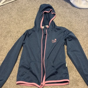 Blå träningsjacka - Snygg blå huvjacka från Play med rosa dragkedja och kantdetaljer. Jackan har en luva och två fickor framtill. Perfekt för att dra över en t shirt när man tränar. Storlek 134-140 för barn. 
