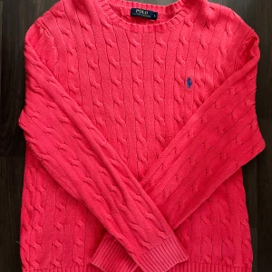 Röd stickad tröja från Ralph Lauren - Säljer en snygg röd stickad tröja från Ralph Lauren med kabelstickat mönster och den klassiska loggan broderad i blått på bröstet. Tröjan har långa ärmar och en rund halsringning. I mycket bra skick.