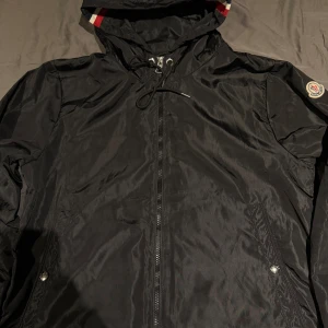 Svart vindjacka från Moncler - Snygg svart vindjacka från Moncler med dragkedja och justerbar huva. Jackan har Moncler-logga på ärmen och detaljer i rött och vitt vid huvan. Perfekt för blåsiga dagar.