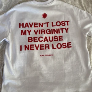 Vit t-shirt från Nude Project - Vit t-shirt från Nude Project med röd text på både fram- och baksida. Framsidan har en liten logotyp och texten 'Winners Club Since Birth'. Baksidan har ett stort tryck med texten 'Haven't lost my virginity because I never lose'. Perfekt för den som vill sticka ut med ett humoristiskt budskap.