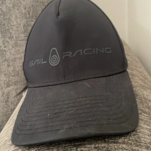 Svart keps från Sail Racing - Snygg svart keps från Sail Racing med justerbart spänne baktill. Perfekt  en dag på sjön. Kepsen har en stilren design med logotyp framtill.