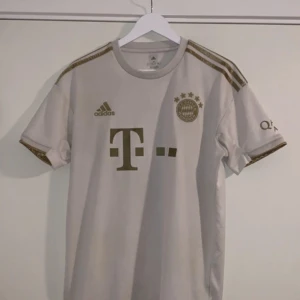 FC Bayern München fotbollströja - Säljer en vit FC Bayern München fotbollströja från Adidas med guldiga detaljer och logotyper. Tröjan har korta ärmar och klubbens emblem på bröstet. Perfekt för fans av laget! ⚽