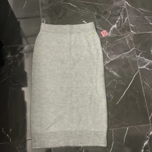 Grå ribbad kjol från Pernilla Wahlgren - Snygg grå ribbad kjol från Pernilla Wahlgren. Kjolen har en stilren design med ett litet guldfärgat ananasmärke som detalj. Perfekt för en chic och bekväm look.