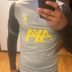 Liverpool träningströja från Nike - Säljer en grå långärmad träningströja från Nike med svarta ärmar och gula detaljer. Tröjan har en dragkedja vid halsen och är perfekt för fotbollsträning. Den har en modern design med klubbmärke och sponsorlogga på bröstet.