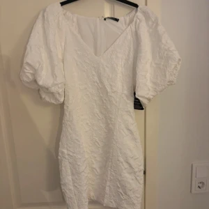 Vit brokadklänning från Gina Tricot - Säljer en elegant vit brokadklänning från Gina Tricot med puffiga kortärmar och en v-ringad design. Klänningen har en dragkedja i ryggen och en vacker textur som ger en lyxig känsla. Perfekt för speciella tillfällen! 🌟