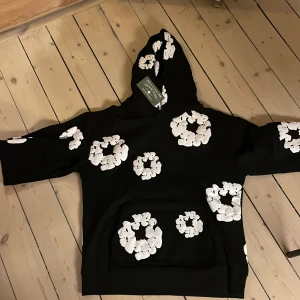 Svart hoodie med vita blommor från Denim Tears - Säljer en svart hoodie från Denim Tears med vita blommor som mönster. Hoodien har en klassisk passform med huva och långa ärmar. använd 3/4 gånger ! Alt org följer med! Pris ej hugget i sten! MVH John