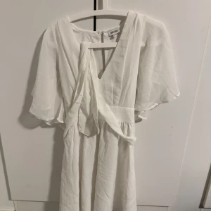 Vit klänning från Bubbleroom - Elegant vit klänning från Bubbleroom med v-ringning och korta ärmar. Klänningen har en fin omlott-detalj och är perfekt för sommarens festligheter. Den är tillverkad i ett lätt och luftigt material som ger en skön känsla.