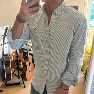 Randig skjorta från Ralph Lauren - Snygg randig skjorta från Ralph Lauren. Skjortan har långa ärmar och en klassisk krage med knappar. Den är dekorerad med det ikoniska broderade logotypen på bröstet. Perfekt för en stilren look!