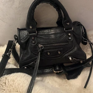 Svart skinn handväska med dragkedja - Snygg svart handväska i skinn med dragkedja och silverdetaljer. Väskan har både handtag och en avtagbar axelrem för flexibilitet. 