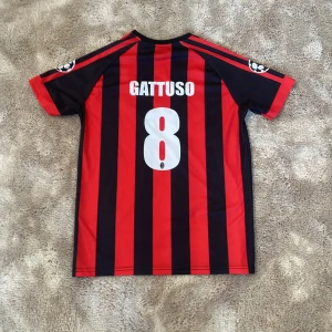 Röd och svart randig Ac Milan fotbollströja, Gattuso - Säljer en klassisk röd och svart randig fotbollströja från Adidas med korta ärmar. Tröjan har ett tryck med 'bwin' på framsidan och 'Gattuso 8' på baksidan. Perfekt för fotbollsfans!