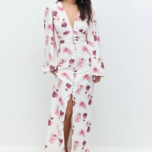 Vit blommig långklänning - Elegant vit långklänning med rosa blommönster och djup v-ringning. Klänningen har långa ärmar och en slits framtill som ger en stilfull touch. Perfekt för sommarens festligheter.
