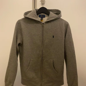 Grå hoodie från Ralph Lauren - Säljer en klassisk grå hoodie från Ralph Lauren med dragkedja och broderad logga på bröstet. Perfekt för en avslappnad stil. Den har en bekväm passform och är tillverkad i mjukt material. NOTREA STORLEK  L  I UNGDOM FRÅN 14/16