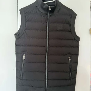 Svart dunväst från Stone Island - Snygg svart dunväst från Stone Island med dragkedja och ribbade kanter. Västen har två praktiska fickor med dragkedja och en broderad logotyp på bröstet. Perfekt för lager-på-lager under kyliga dagar. Använda även under sommardagarna.