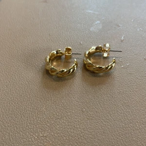 Guldiga örhängen i tvinnad design - Säljer ett par eleganta guldiga örhängen med en tvinnad design. Perfekta för att ge en stilren touch till din outfit. Köp direkt eller ge prisförslag!