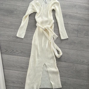 Vit ribbad klänning från Fashionkilla - Snygg vit ribbad klänning från Fashionkilla med lång ärm och v-ringning. Klänningen har en knytning i midjan och en slits framtill som ger en elegant touch. Perfekt för en stilren look.