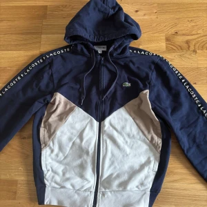 Blå och beige hoodie från Lacoste - Storlek S passar M också! Säljer en asfet zip hoodie från Lacoste i blått  beige och vitt med dragkedja och huva. Tröjan har Lacoste-logga på bröstet och text längs ärmarna. Perfekt för en casual look. Inga defekter och bra skick!