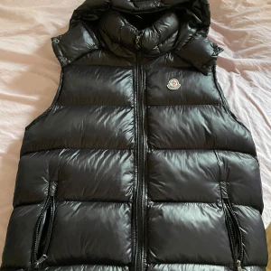 Svart dunväst från Moncler - Säljer en snygg svart dunväst från Moncler med dragkedja och huva. Västen har ett broderat Moncler-märke på bröstet och praktiska fickor med dragkedjor. Perfekt för kyliga dagar! Nfc finns på logan, helt ny,storlek 4 (L)  alla detaljer finns med pris kan diskuteras 