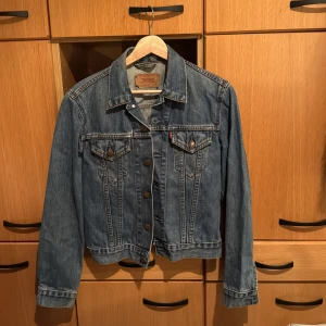 Blå jeansjacka från Levi's - Säljer en klassisk blå jeansjacka från Levi's i storlek S. Jackan har långa ärmar och knappar framtill samt två bröstfickor med knappstängning. Perfekt för en avslappnad stil.