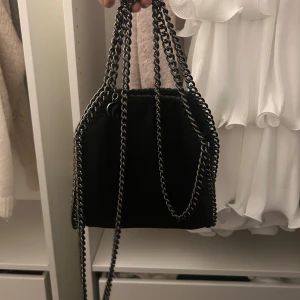 Svart axelväska med kedjedetaljer - Snygg svart axelväska med kedjedetaljer som ger en cool look. Väskan har en stilren design och passar perfekt för att lyfta din outfit. Den är lätt att bära med sina kedjehandtag och har en praktisk storlek för dina nödvändigheter.