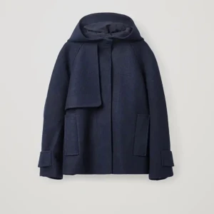 Mörkblå kappa med huva från COS - Väldigt snygg och bekväm mörkblå kappa från COS med huva och långa ärmar. Jackan har en stilren design med dolda knappar och stora fickor framtill. 100% Ull.