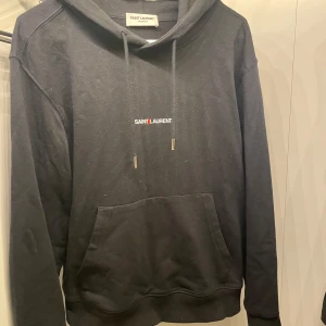 Svart hoodie från Saint Laurent - Snygg svart hoodie från Saint Laurent med logotyp framtill. Den har en klassisk design med justerbar huva och en stor magficka. Perfekt för en stilren och avslappnad look. Inga deffekter, behöver ba tvättas.  Använd tre gånger. Skriv vid till mig för mer bilder eller frågor🤩💫