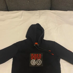 Svart hoodie från Hugo - Säljer en stilren svart hoodie från Hugo med en stor logotyp i rött och vitt på bröstet. Hoodien har en justerbar huva med svarta snören och röda detaljer. Perfekt för en avslappnad stil.