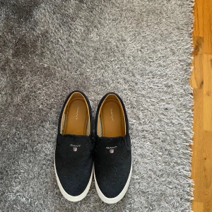 Svarta slip-on sneakers från Gant - Snygga svarta slip-on sneakers från Gant med vit sula och rund tå. Skorna har en diskret Gant-logga på ovansidan och är perfekta för en avslappnad stil. De är lätta att ta på och av. Perfekta nu till sommaren. Passar mig utmärkt som har 42/5