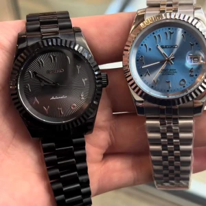 Två Seiko-klockor i svart och silver - Två eleganta Seiko-klockor, en i helsvart med automatisk urverk och en i silver med blå urtavla och datumvisning. Båda har arabiska siffror och räfflade boetter. Perfekta för den stilmedvetna.