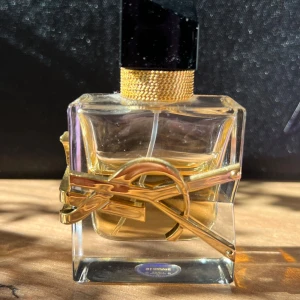 Yves Saint Laurent Libre Eau de Parfum 30 ml - En lyxig parfymflaska från Yves Saint Laurent med guldiga detaljer och svart kork. Flaskan har en unik design med ett stort YSL-logotypband i metall runt glaset och innehåller en gyllene vätska. Säljes då jag har blivit allergisk mot dofter.  Ungefär 18 ml kvar därav 380 kr (nypris 749 kr). 
