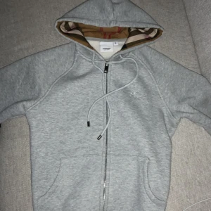 Grå hoodie med dragkedja från Burberry - Säljer en grå hoodie från Burberry med dragkedja och huva. Insidan av huvan har det klassiska Burberry-mönstret i beige, rött och svart. Tröjan har fickor framtill och justerbar dragsko vid huvan.