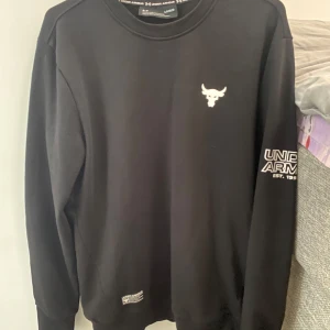 Svart sweatshirt från Under Armour Project Rock - Svart sweatshirt från Under Armour Project Rock med logga på bröstet och tryck på ärmen. Klassisk rund halsringning och långa ärmar. Perfekt för träning eller chill. Materialet känns mjukt och bekvämt. Storlek L men känns mer som M i längd och midja. 
