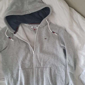 Grå hoodie med dragkedja från Tommy Hilfiger Denim - Säljer en ljusgrå hoodie från Tommy Hilfiger Denim med dragkedja och huva. Tröjan har vita snören med röda och blå detaljer, två fickor fram och liten broderad logga på bröstet. Skriv vid frågor!🥰