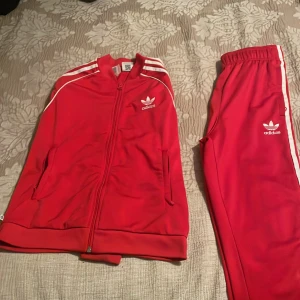 Röda Adidas mjukisbyxor med vita ränder - Säljer ett par klassiska röda Adidas mjukisbyxor med vita ränder längs sidorna och broderad logga framtill. Byxorna har resår i midjan och är perfekta för en sportig och avslappnad stil.