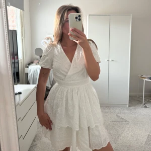 Vit kortklänning med broderade blommor och v-ringning - Superfin vit klänning med broderade blommor, v-ringning och omlottliv. Puffärmar och markerad midja, volang i kjolen och öppen detalj i ryggen. Perfekt för sommarens alla tillfällen! Klänningen är använt bara en gång men är tyvärr för liten för mig.