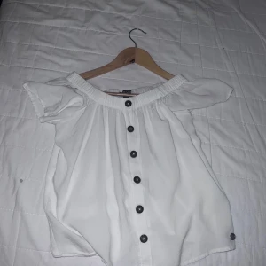 Vit offshoulder blus med knappar - Säljer en söt vit offshoulder blus med svarta knappar framtill. Blusen har korta ärmar och en bred resår vid axlarna som ger en snygg passform. Perfekt till sommaren!