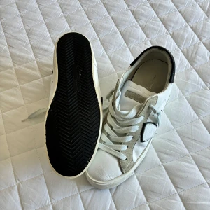 Vita sneakers från Philippe Model - Säljer ett par vita sneakers från Philippe Model med gråa och svarta detaljer. Skorna har snörning, rund tå och platt sula. Perfekta för en stilren och avslappnad look.