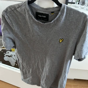 Grå t-shirt från Lyle & Scott - Säljer en klassisk grå t-shirt från Lyle & Scott med rund halsringning och den ikoniska gula loggan broderad på bröstet. Perfekt basic-plagg i mjuk bomull som passar till det mesta.