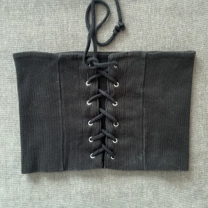 Svart ribbad tubtopp med snörning - Svart ribbad tubtopp med snörning framtill och metallöljetter. Toppens material är stretchigt och sitter tight runt kroppen. Perfekt till fest! Hör av dig för mer info 🤗