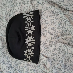 Svart mössa med vitt mönster från Everest - Svart mössa från Everest med vitt, pixelinspirerat mönster längs kanten. Tillverkad i stretchigt syntetmaterial som passar de flesta huvudstorlekar. Perfekt för kalla dagar och enkel att matcha med vinterjackan.