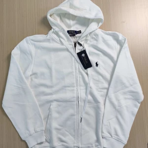Vit hoodie från Polo Ralph Lauren - Säljer en klassisk vit hoodie från Polo Ralph Lauren i storlek M. Tröjan har dragkedja framtill, huva med snörning och två fickor. Obs snörerna är ej på och de finns en liten färg fläck på baksidan som enkelt kan tas bort