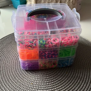 Låda med färgglada gummiband och tillbehör för armbandstillverkning - Säljer en transparent plastlåda med flera fack fyllda med färgglada gummiband i rosa, grönt, blått, orange, lila och flerfärgade varianter. Medföljer rosa vävram, krokar och clips för att skapa egna armband. Perfekt för pyssel och kreativt skapande.
