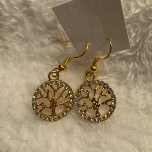 Guldiga örhängen med livets träd och strass - Snygga guldiga örhängen med rund berlock i form av livets träd. Runt kanten sitter glittrande stenar som ger extra bling. Perfekta för att lyfta din look med en elegant touch. Krokarna är i sterling silver 