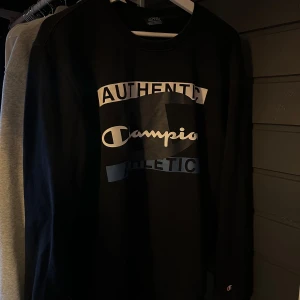 Svart sweatshirt från Champion - Säljer en svart sweatshirt från Champion med tryckt text 'Authentic Champion Athletic' i vitt och blått på bröstet. Tröjan har rund halsringning och långa ärmar. Perfekt för en sportig och avslappnad stil.
