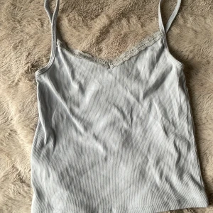 Ljusblå ribbad linnetopp med spets från H&M - Säljer en ljusblå ribbad linnetopp från H&M med smala axelband och vacker spetskant vid urringningen. Perfekt för sommaren och enkel att matcha med olika outfits.